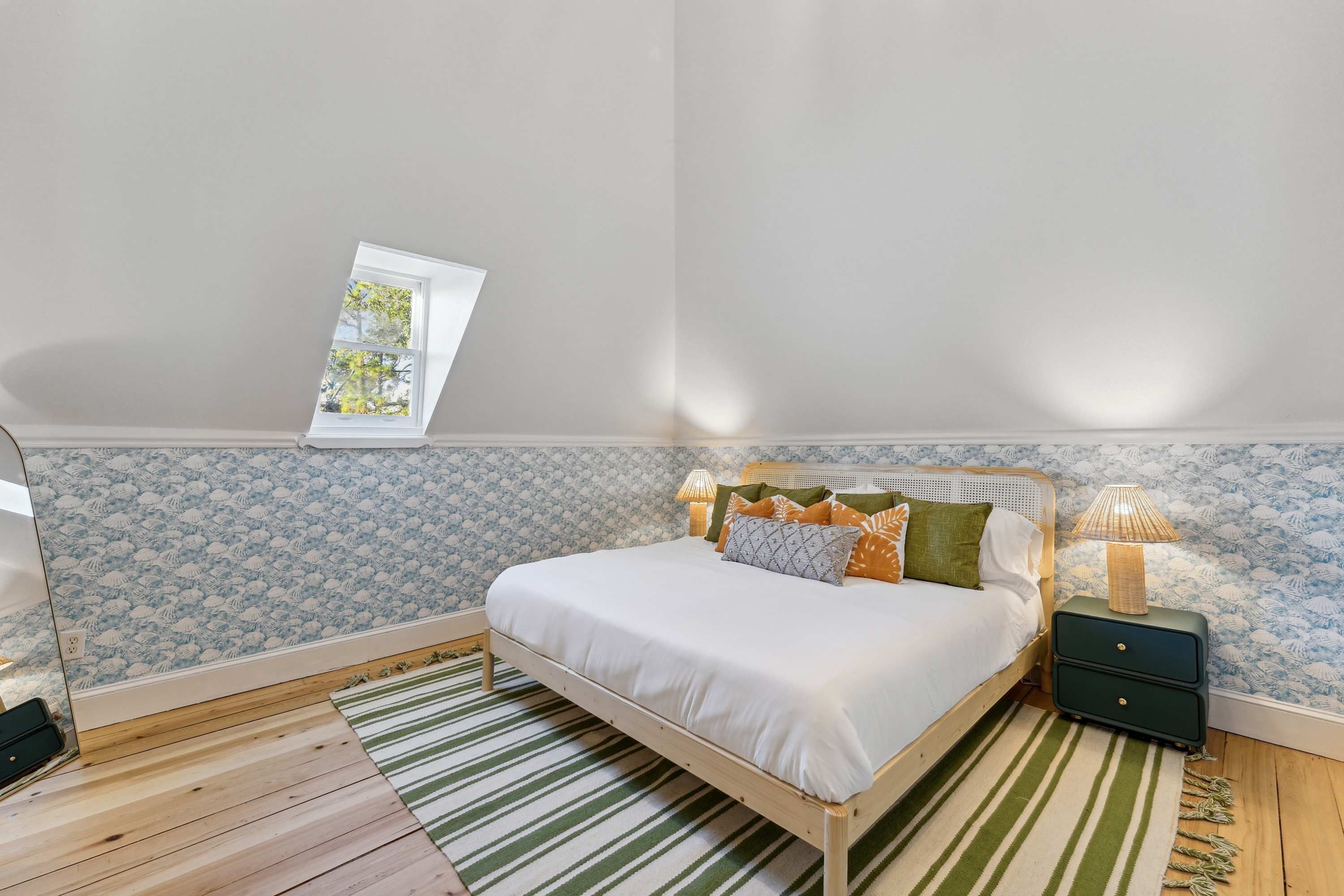 Une chambre à coucher avec un lit, une table de chevet, une lampe, une fenêtre et un mur décoré d’un motif.