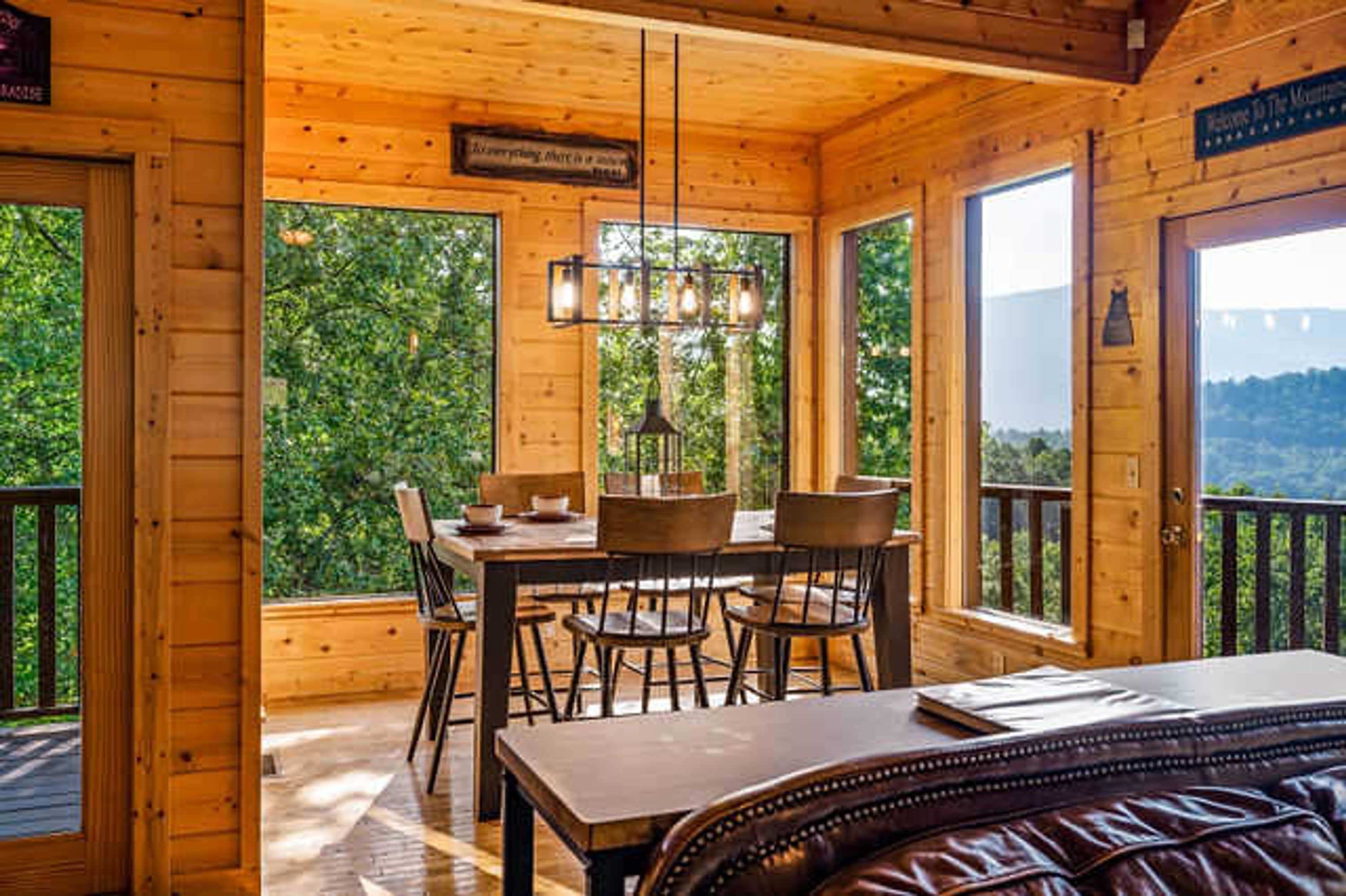 A wooden cabin with a dining area, a bed, and large windows overlooking a forested landscape.