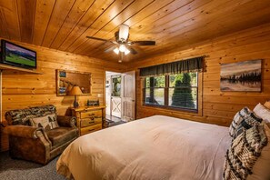 A wooden cabin bedroom with a bed, a desk, a chair, a television, and a window with a view of trees. - Private Mtn Views Movie Theater Hot Tub Pool Table (Sevierville)