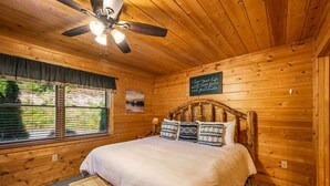 Ein Schlafzimmer einer Holzhütte mit einem Bett, einem Deckenventilator, einem Fenster mit Jalousien und einer kleinen Holzbank.