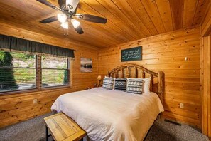 Ein Schlafzimmer einer Holzhütte mit einem Bett, einem Deckenventilator, einem Fenster mit Jalousien und einer kleinen Holzbank.