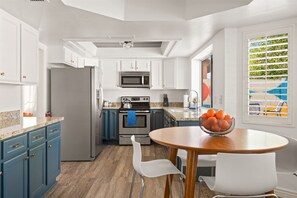 Eine moderne Küche mit weißen Schränken, einem Edelstahlkühlschrank, einer Mikrowelle und einem Backofen. Ein Esstisch mit einer Obstschale und zwei weißen Stühlen steht daneben.