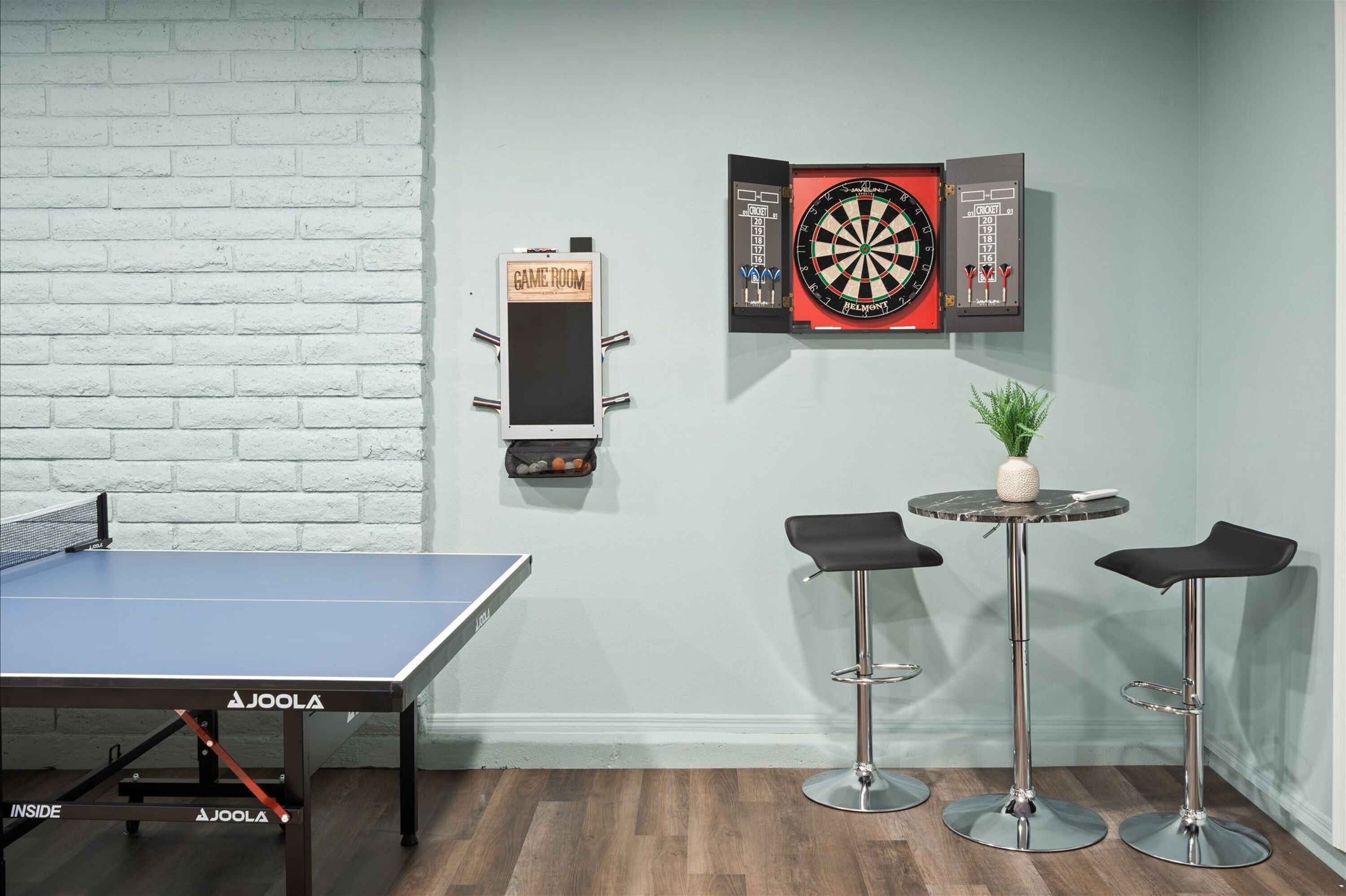 Une pièce avec une table de ping-pong, deux tabourets de bar, un jeu de fléchettes et une petite plante.