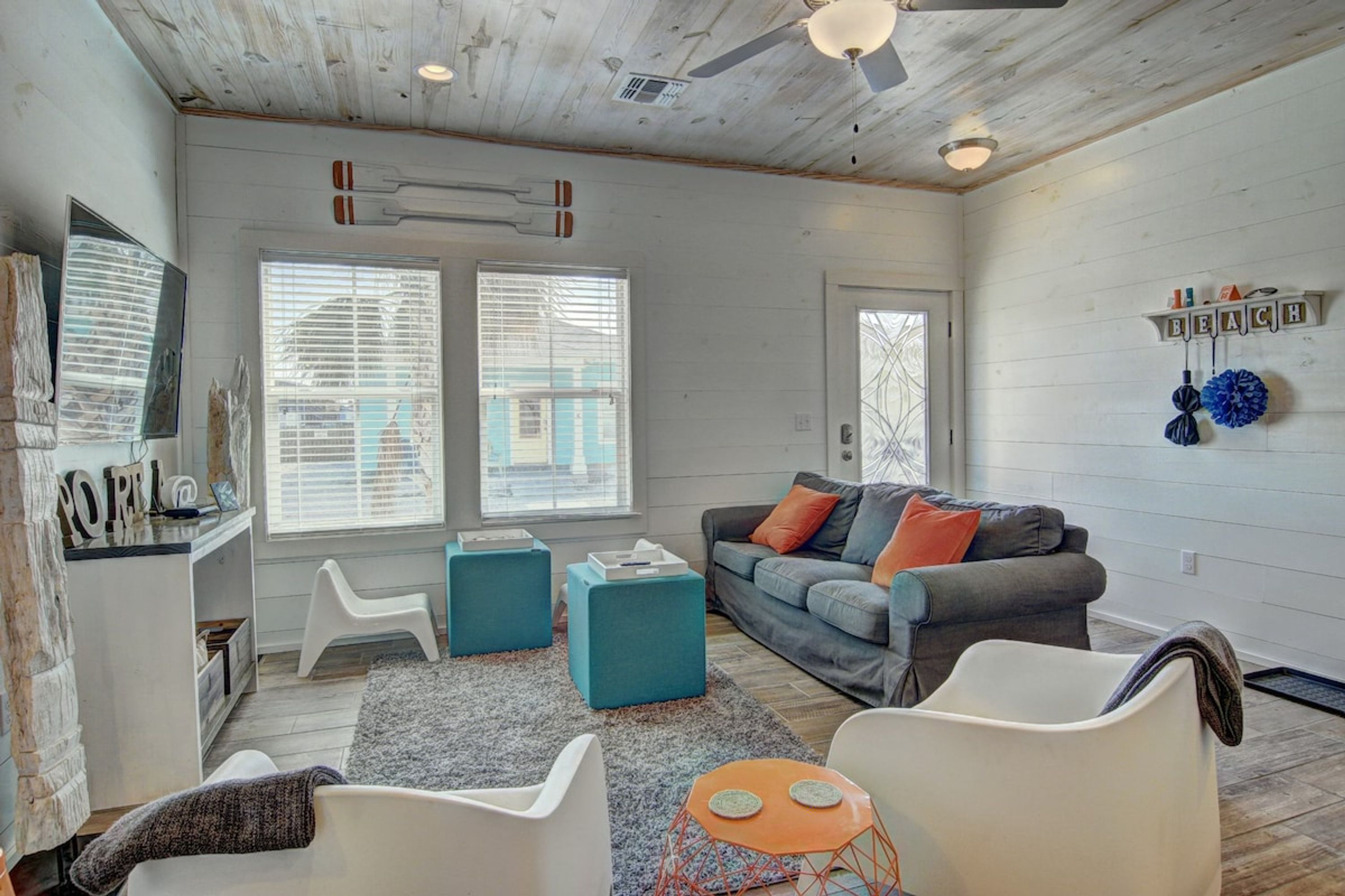 A spacious living room with a gray sofa, teal ottomans, and a white armchair. There are two windows with blinds, a ceiling fan, and a wall-mounted shelf with decorative items.