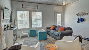 A spacious living room with a gray sofa, teal ottomans, and a white armchair. There are two windows with blinds, a ceiling fan, and a wall-mounted shelf with decorative items. - Beach Please- Family Friendly Community Pool (Port Aransas)