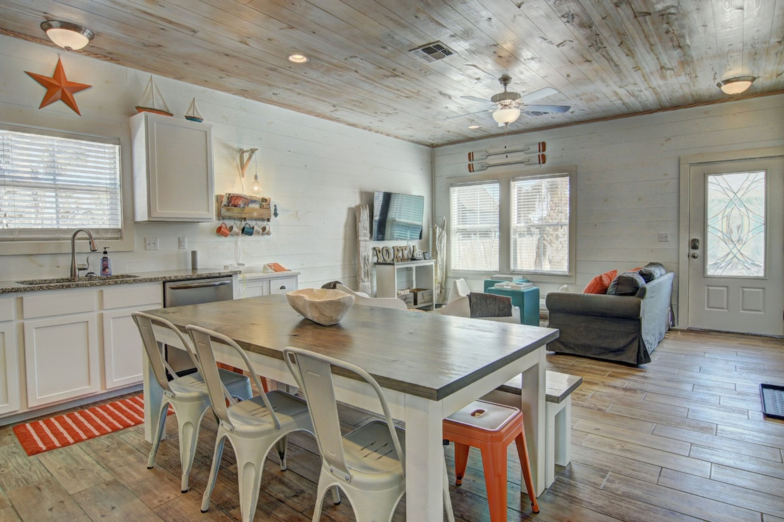 A spacious kitchen with a wooden table and white chairs, a dining area with a sofa, and a large window with blinds.