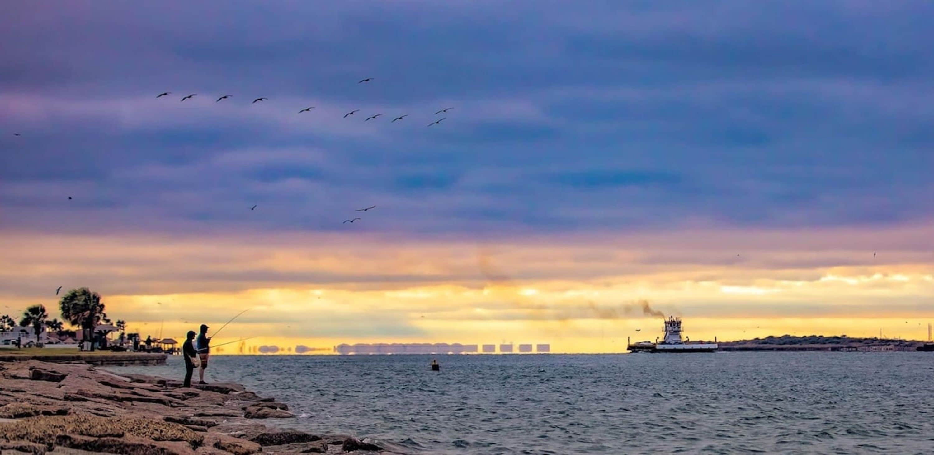 Escena costera con un litoral rocoso, un barco a lo lejos y un cielo al atardecer.