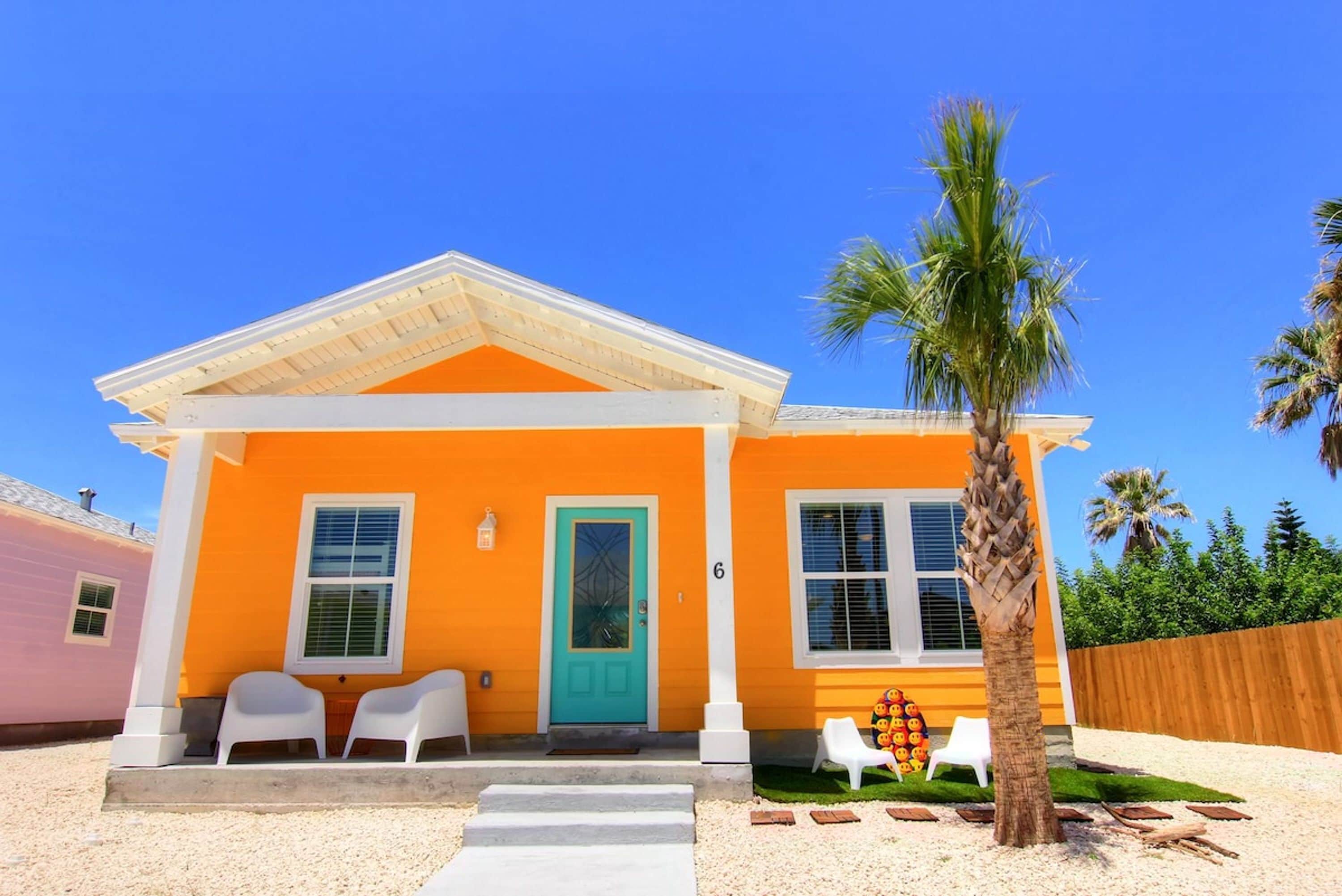 A two-story house with a bright orange exterior, a turquoise door, and a small porch with white chairs.