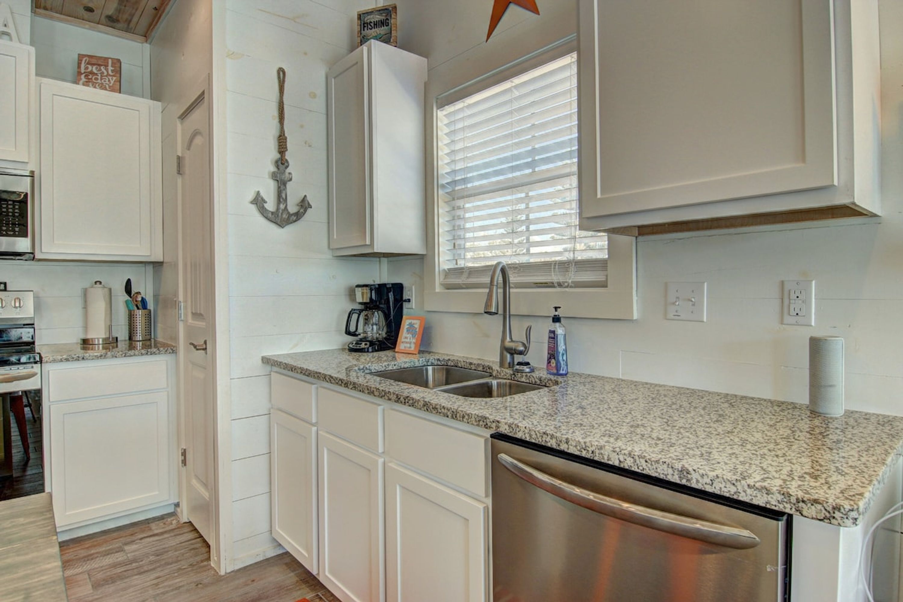 A kitchen with white cabinets, a stainless steel dishwasher, a coffee maker, and a window with blinds.
