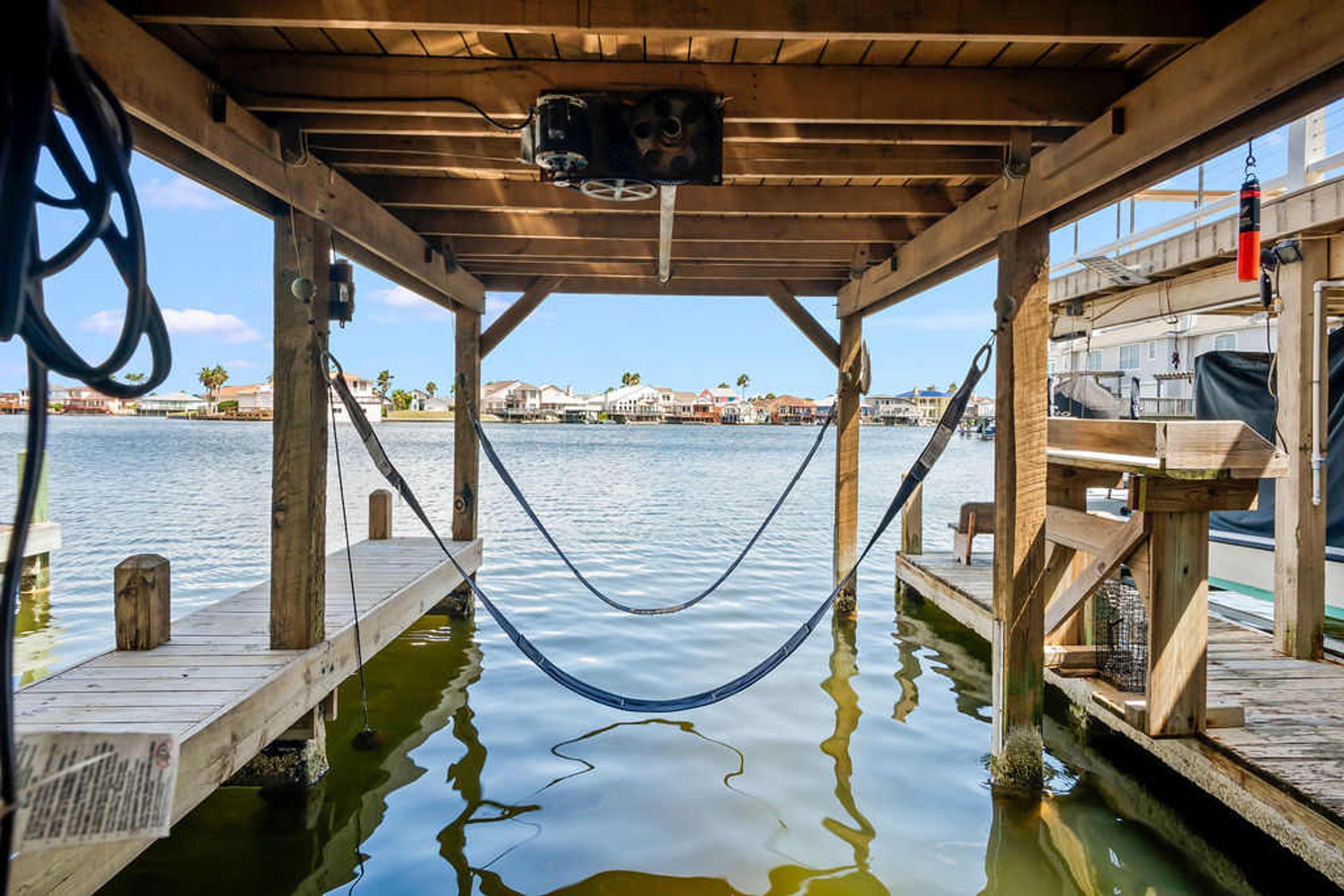 Un muelle de madera con una hamaca, una estructura de madera y vistas a un cuerpo de agua tranquilo con edificios al fondo.
