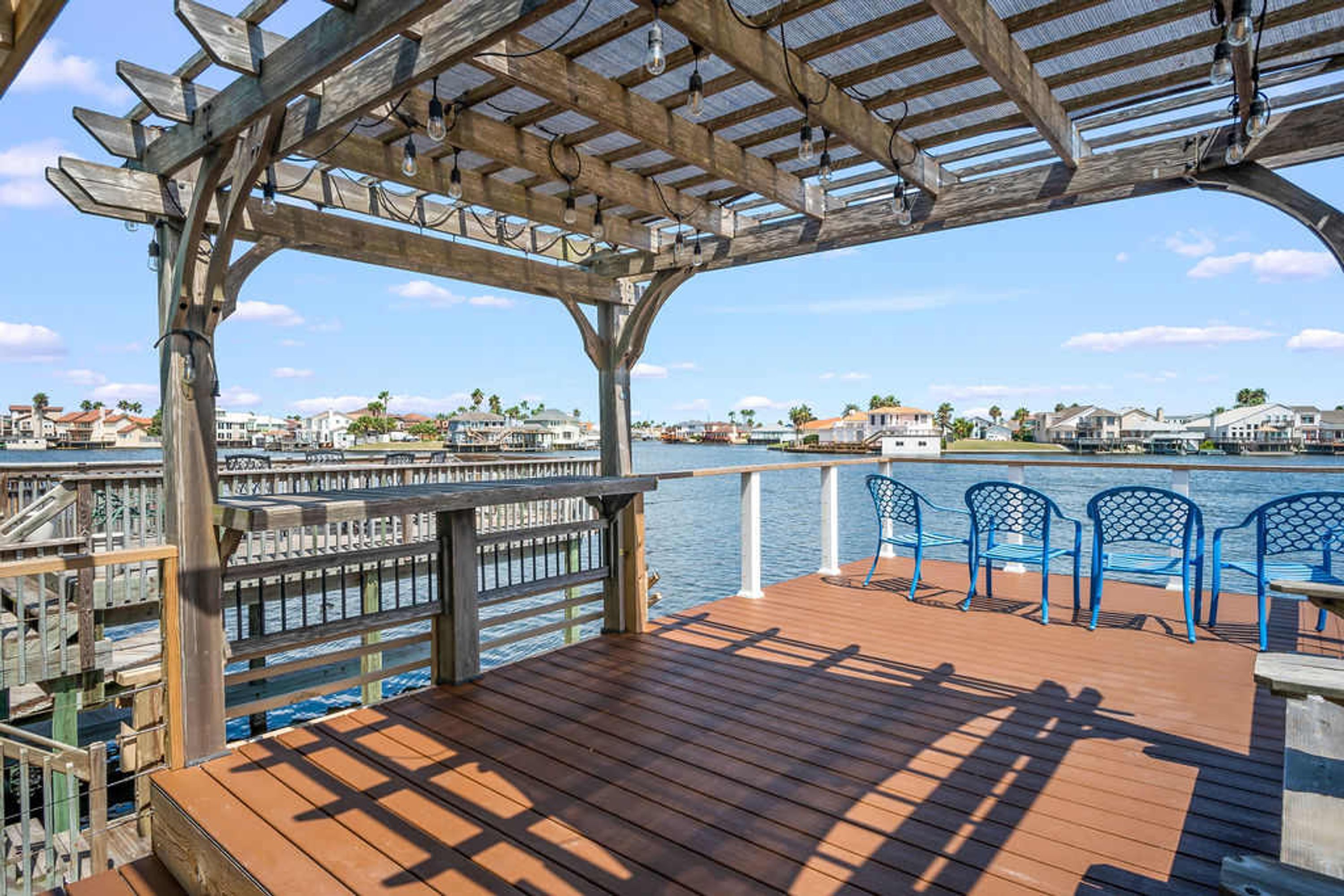 Una terraza de madera con un pérgola, con vista a un frente marítimo con casas y un cielo despejado.