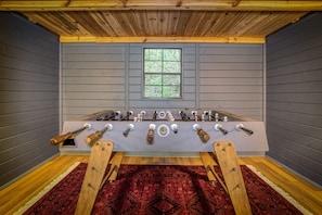 A room with a foosball table, a window with a view of trees, and a patterned rug on the floor. - Blue Ridge Treehouse Escape Views Hot Tub (Blue Ridge)