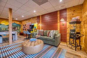 Una habitación acogedora con un sofá verde, un otomanillo redondo y una mesa de centro. Hay dos sillones, una lámpara y una pequeña mesa con lámpara. Las paredes están decoradas con murales coloridos y un diseño de paneles de madera.