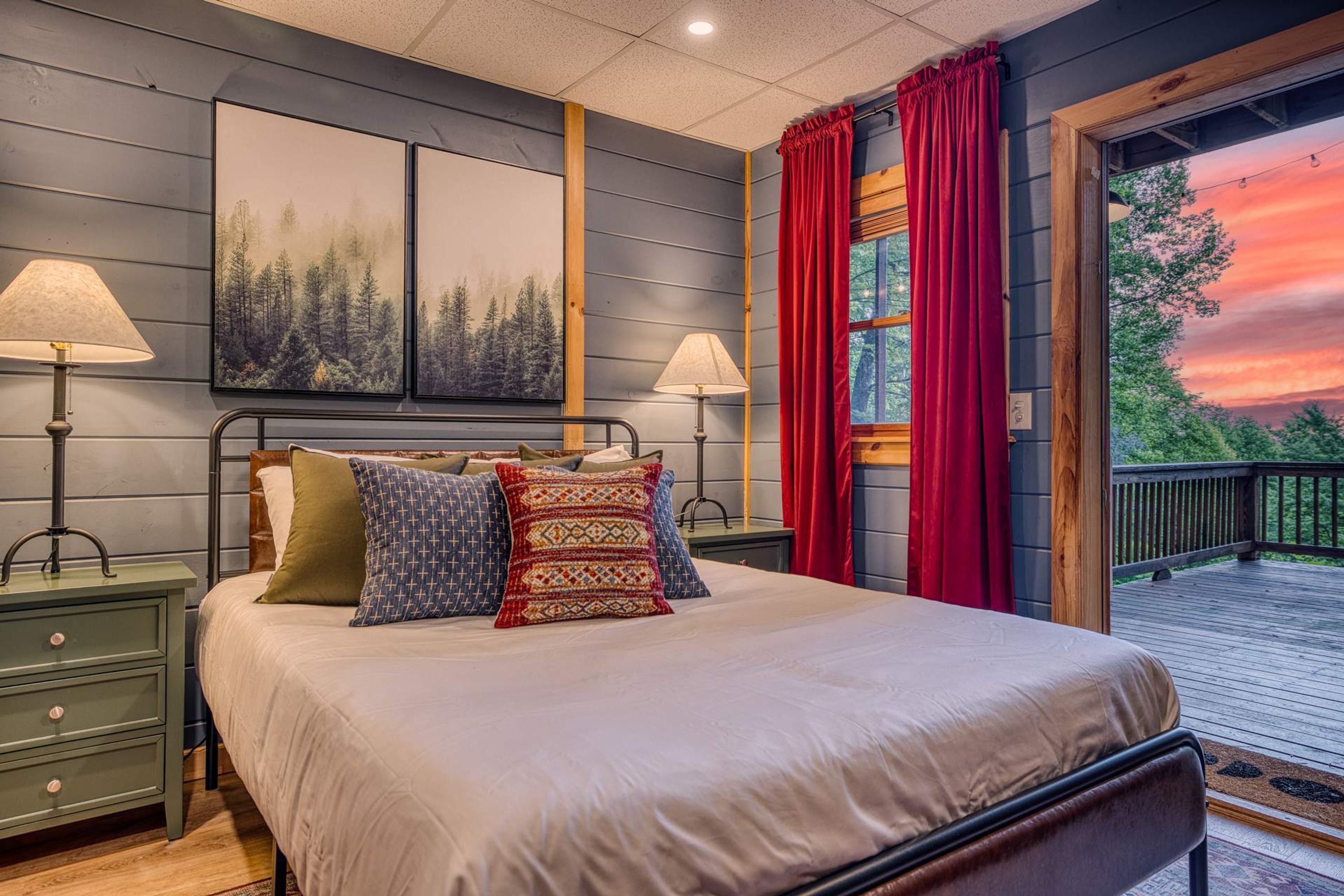 A bedroom with a bed, two nightstands, a window with red curtains, and a view of trees.