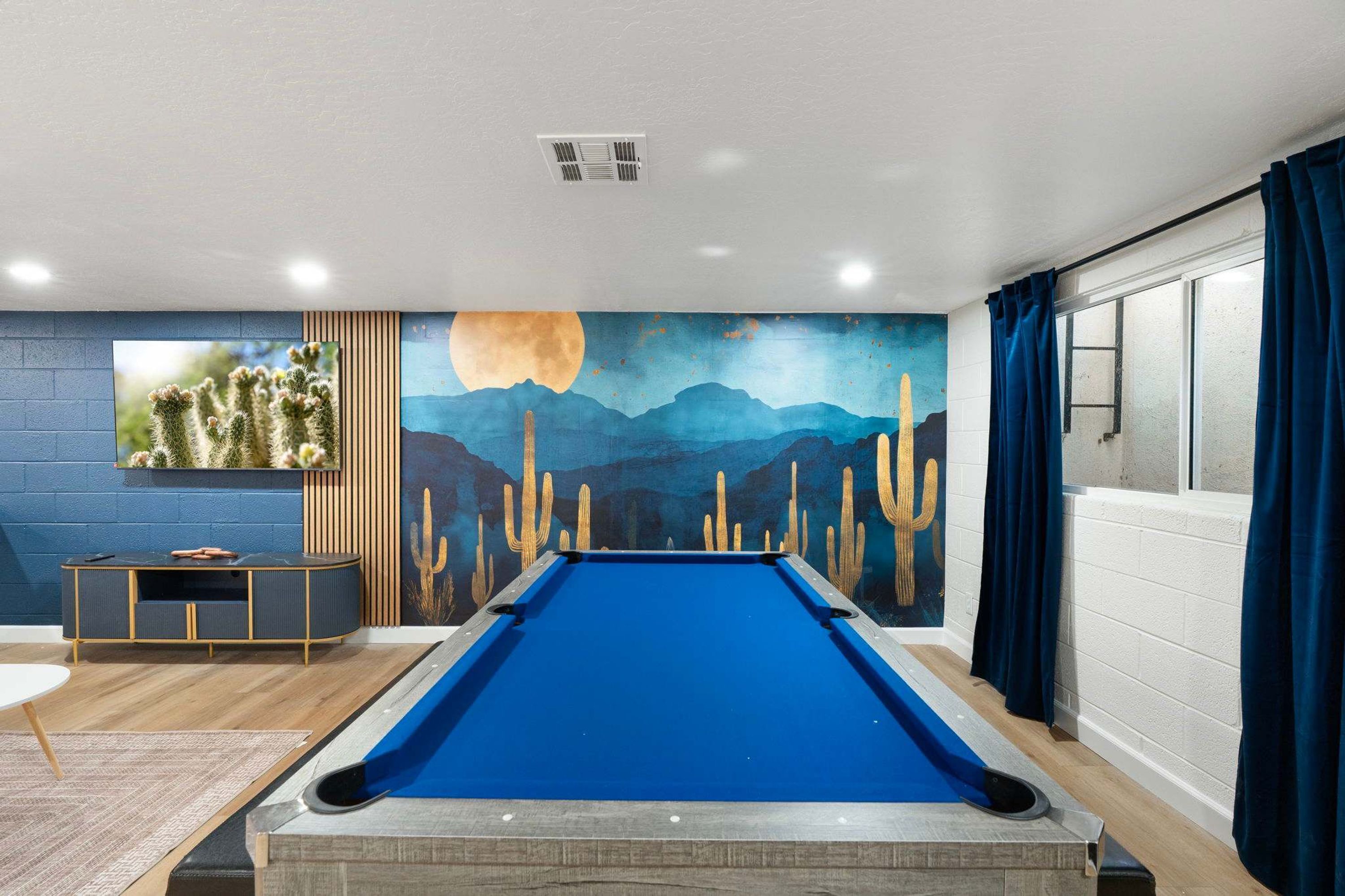 A pool table in a room with a mural of cacti and a moon, a blue wall, and a window with curtains.