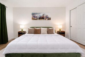 Ein Schlafzimmer mit einem großen Bett, zwei Nachttischen mit Lampen, einem grünen Kopfteil und einer Tür auf der rechten Seite.
