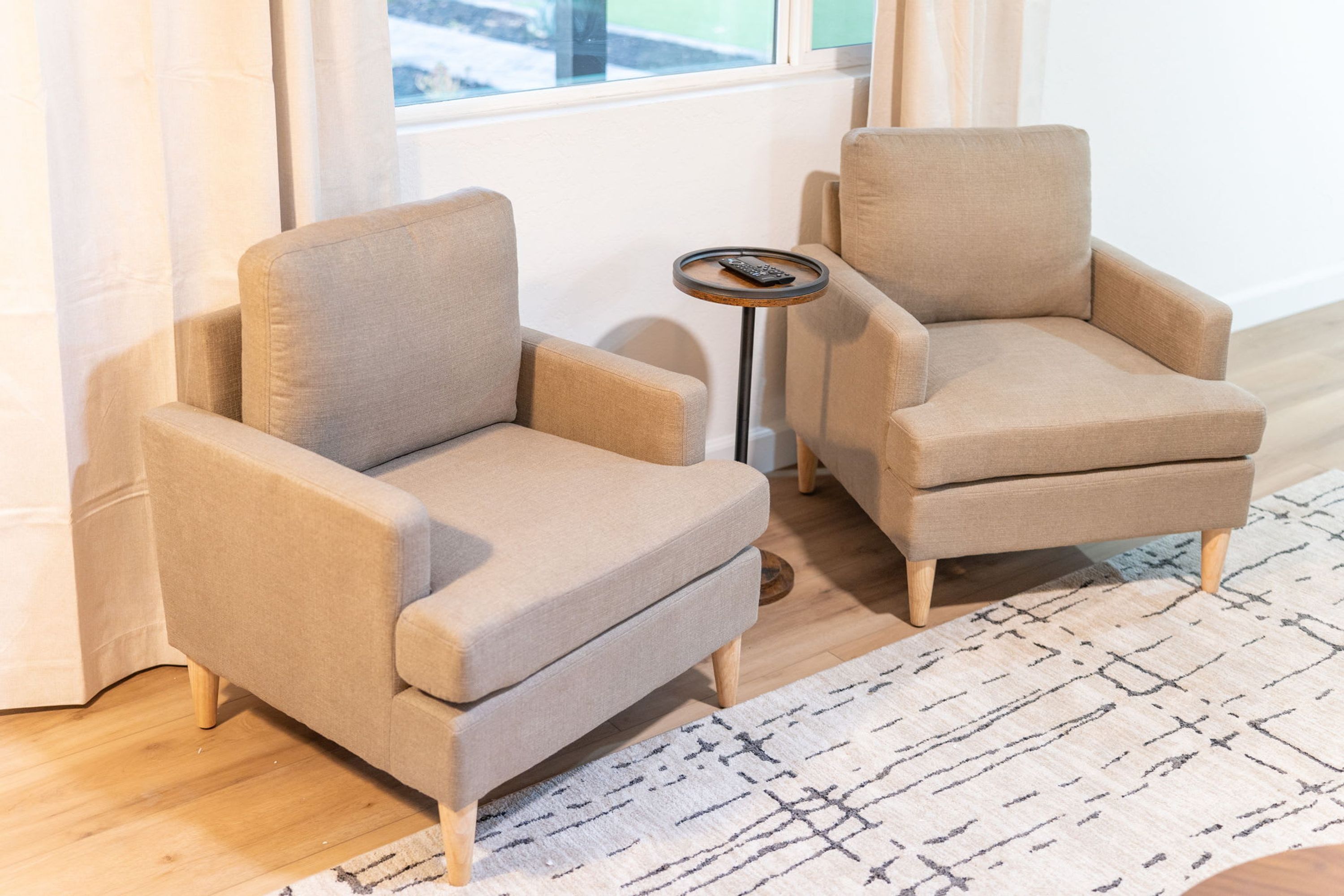 Une pièce avec deux fauteuils beiges, une petite table ronde avec une télécommande, et un tapis à motifs posé sur un plancher en bois.