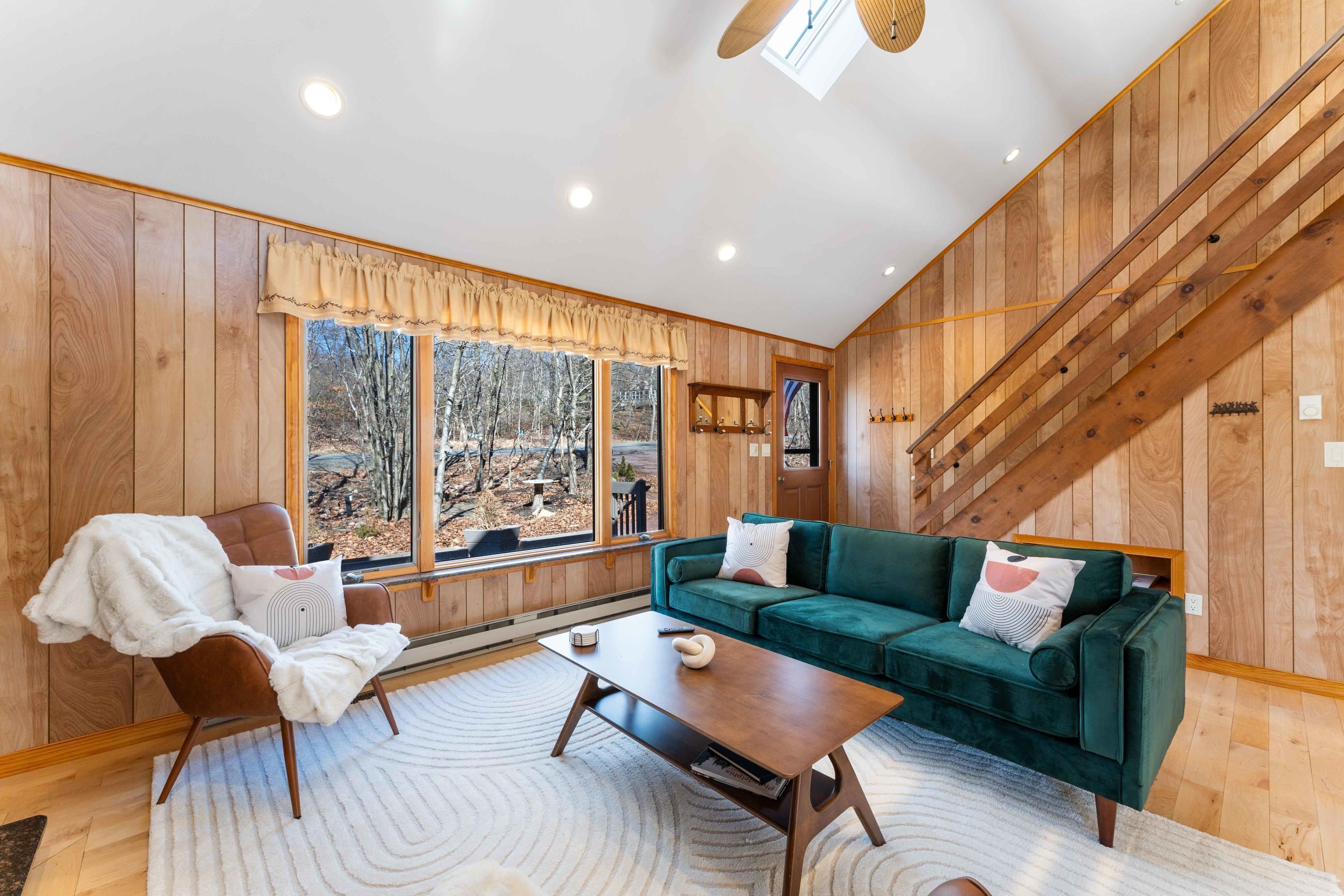 A cozy living room with a green sofa, a wooden table, and a chair. There is a large window with a view of trees and a wooden staircase.