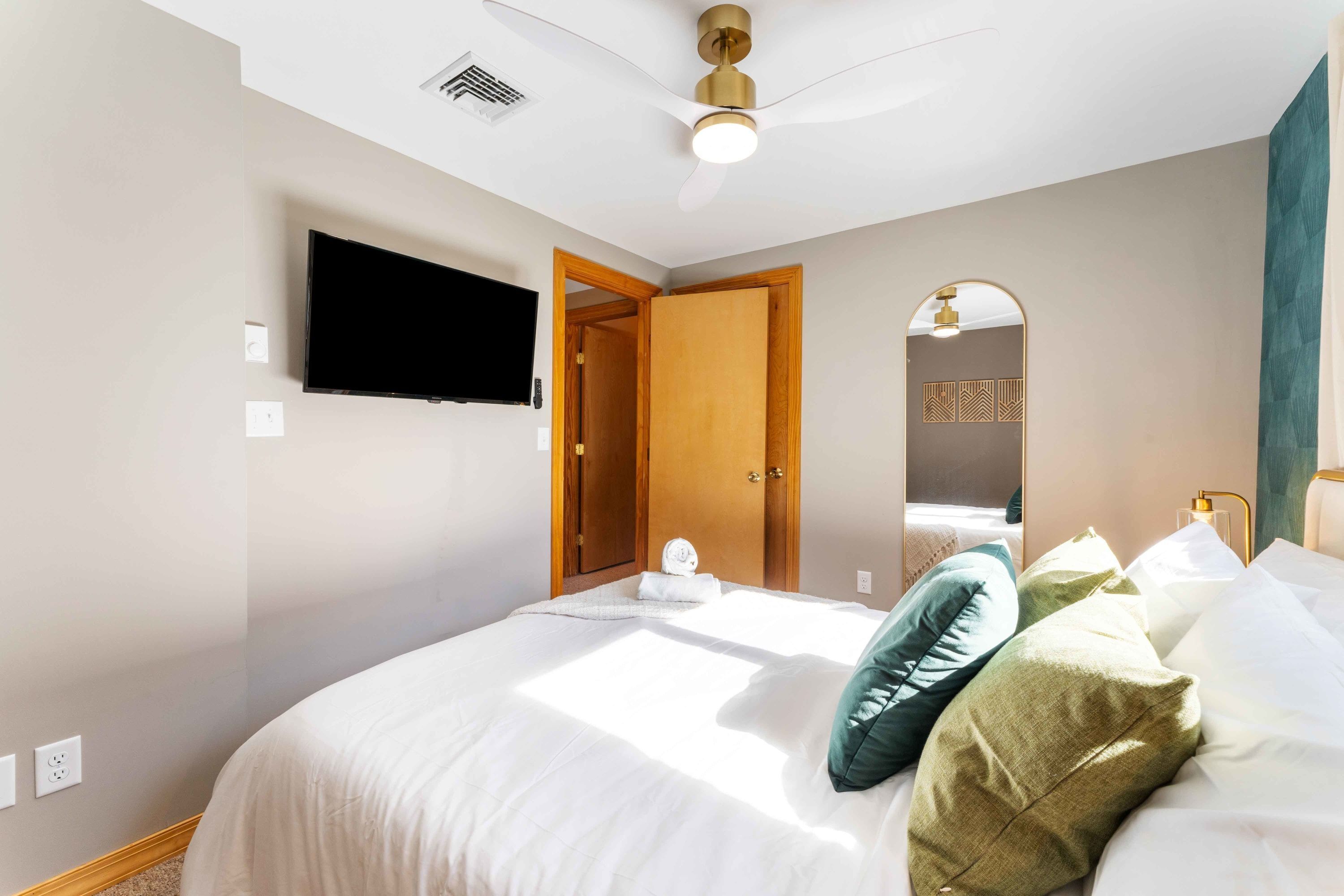 A hotel room with a bed, a flat-screen TV mounted on the wall, a ceiling fan, and a door leading to a bathroom.