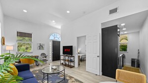 A modern living room with a sofa, a coffee table, and a TV. There are windows with blinds, a door, and a kitchen area visible. - Pineapple Paradise 1 13 min to Beach Fire Pit (Sarasota)