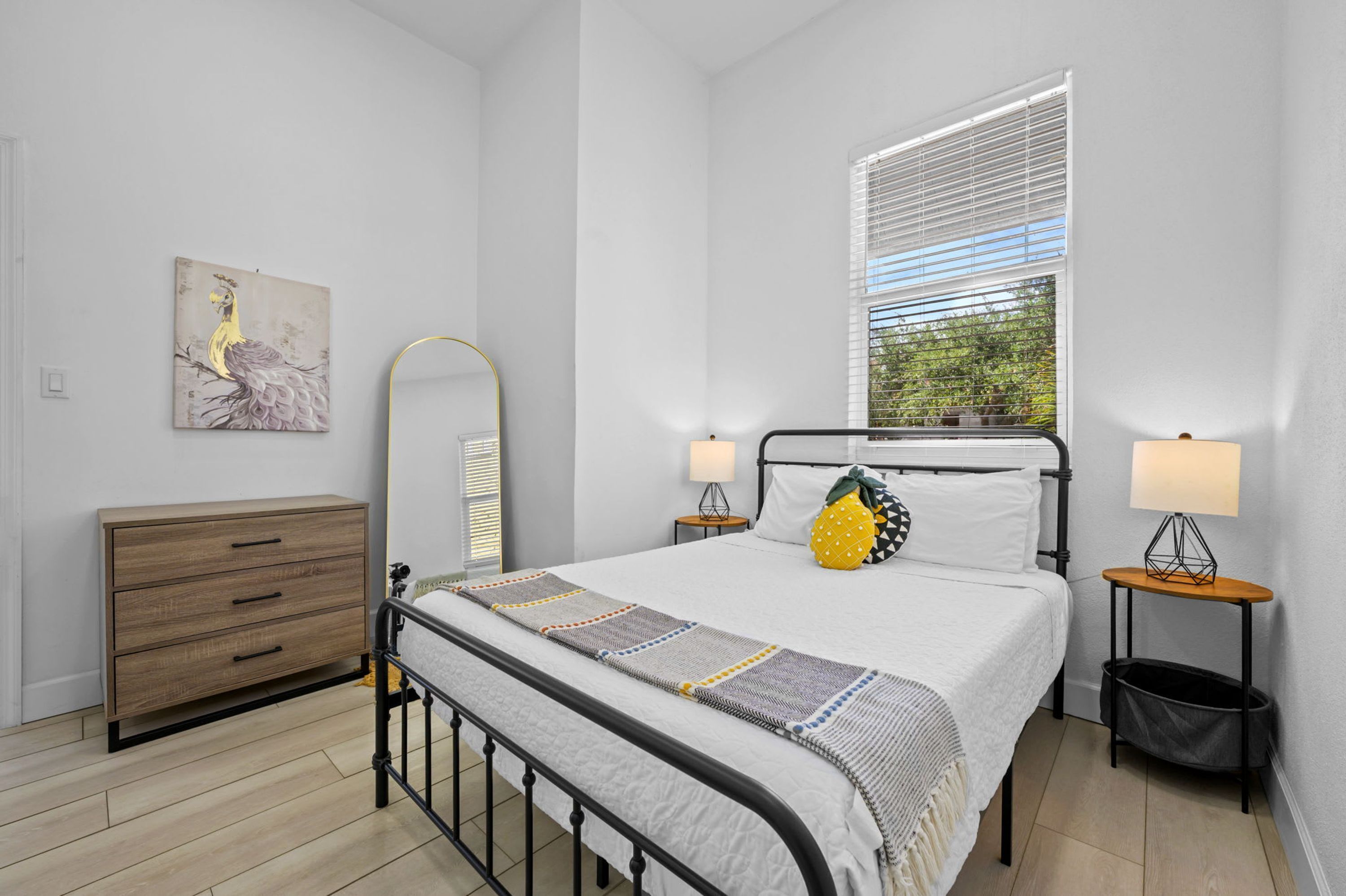 A bedroom with a bed, bedside lamps, a dresser, a mirror, and a window with blinds.