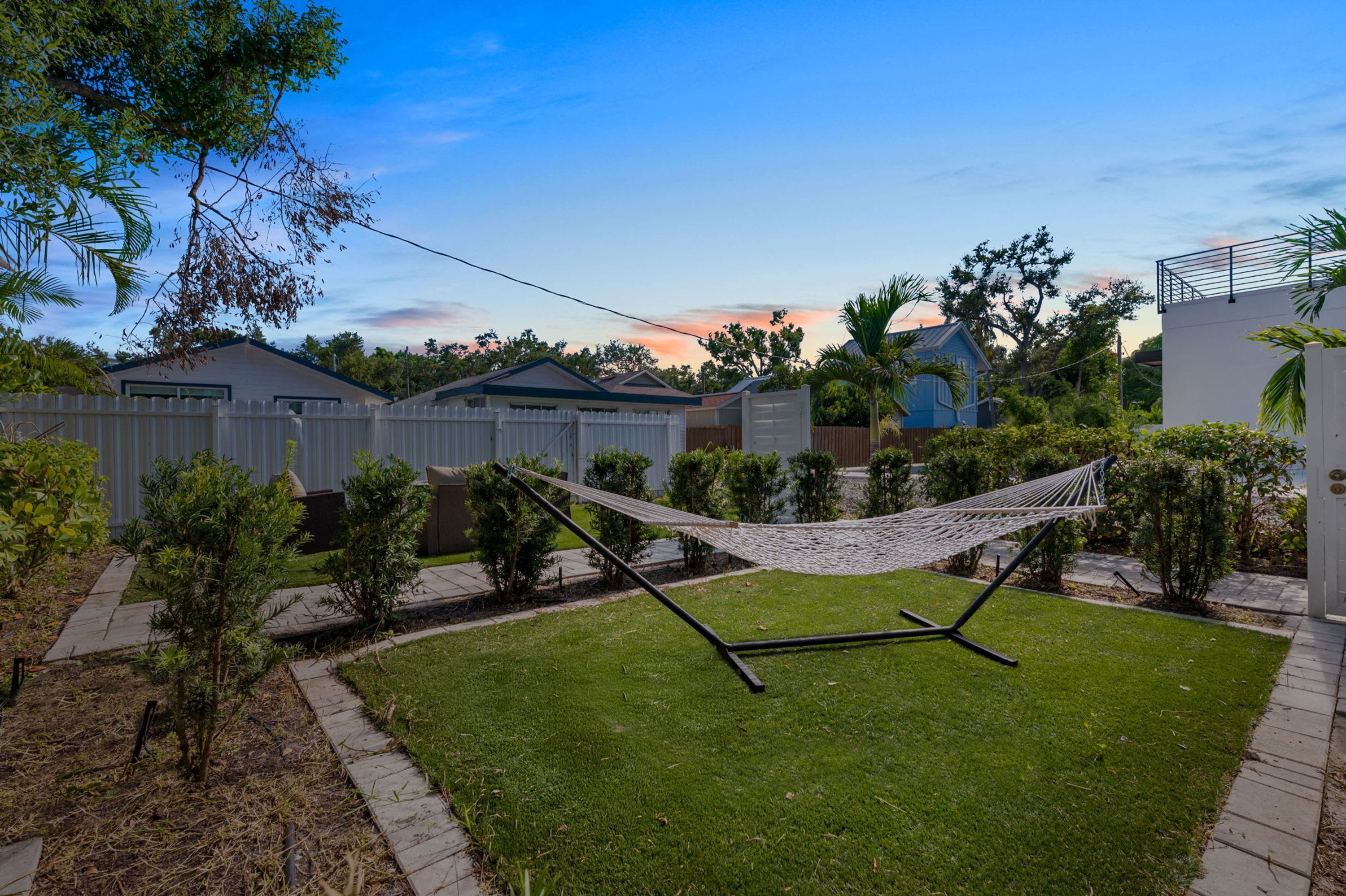 A backyard with a hammock, a lawn, a white fence, and houses in the background.