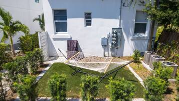A residential backyard with a hammock, a small lawn, and a white building with windows and outdoor electrical boxes.
