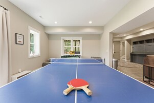 A ping pong table with a single paddle in the foreground, a window with a view of trees, and a door leading to a garage.
