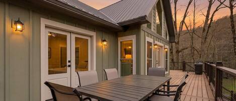 A wooden deck with a dining table and chairs, a covered patio area, and a view of trees and hills.