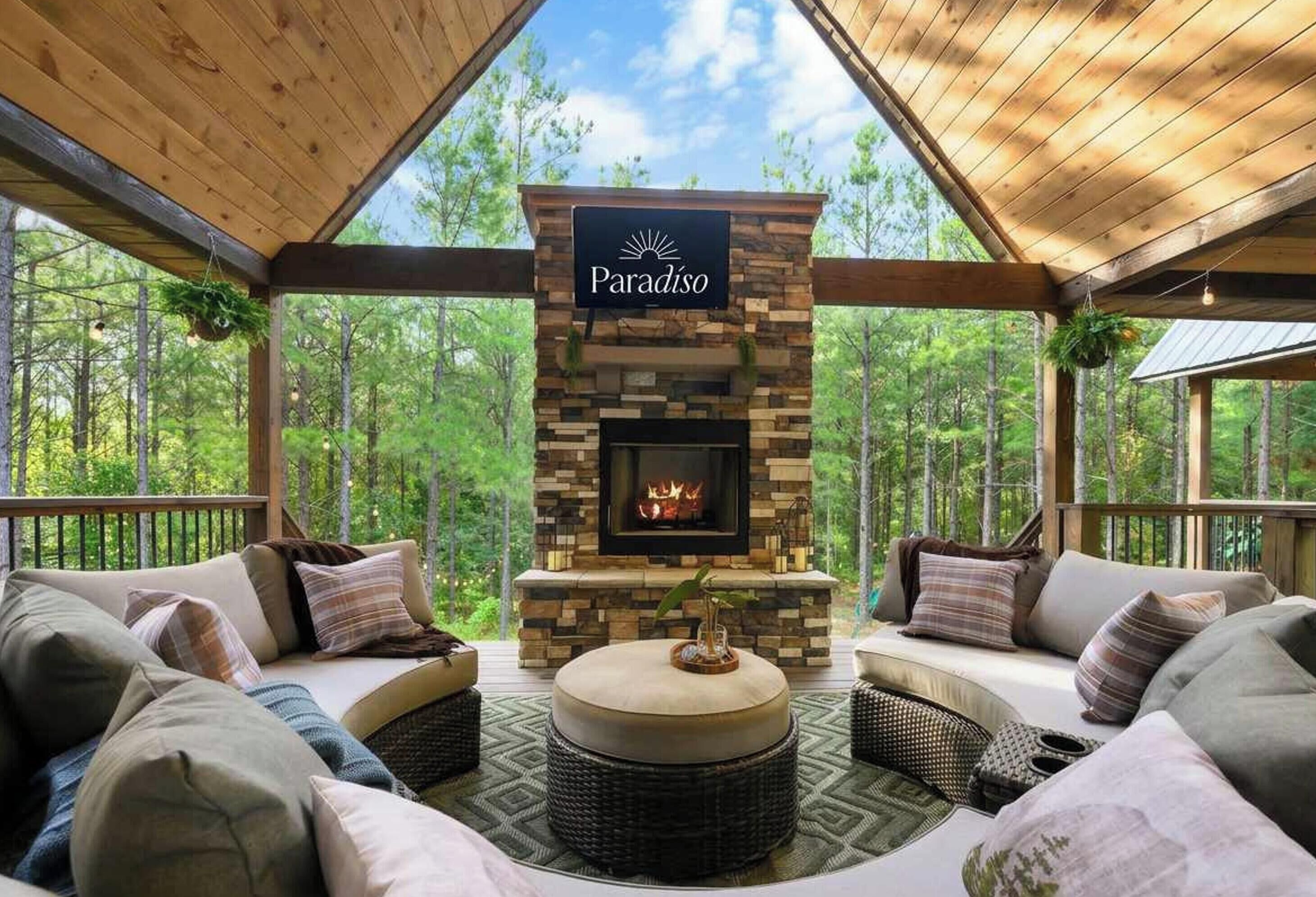 A cozy outdoor seating area with a stone fireplace, surrounded by wooden beams and overlooking a forest.