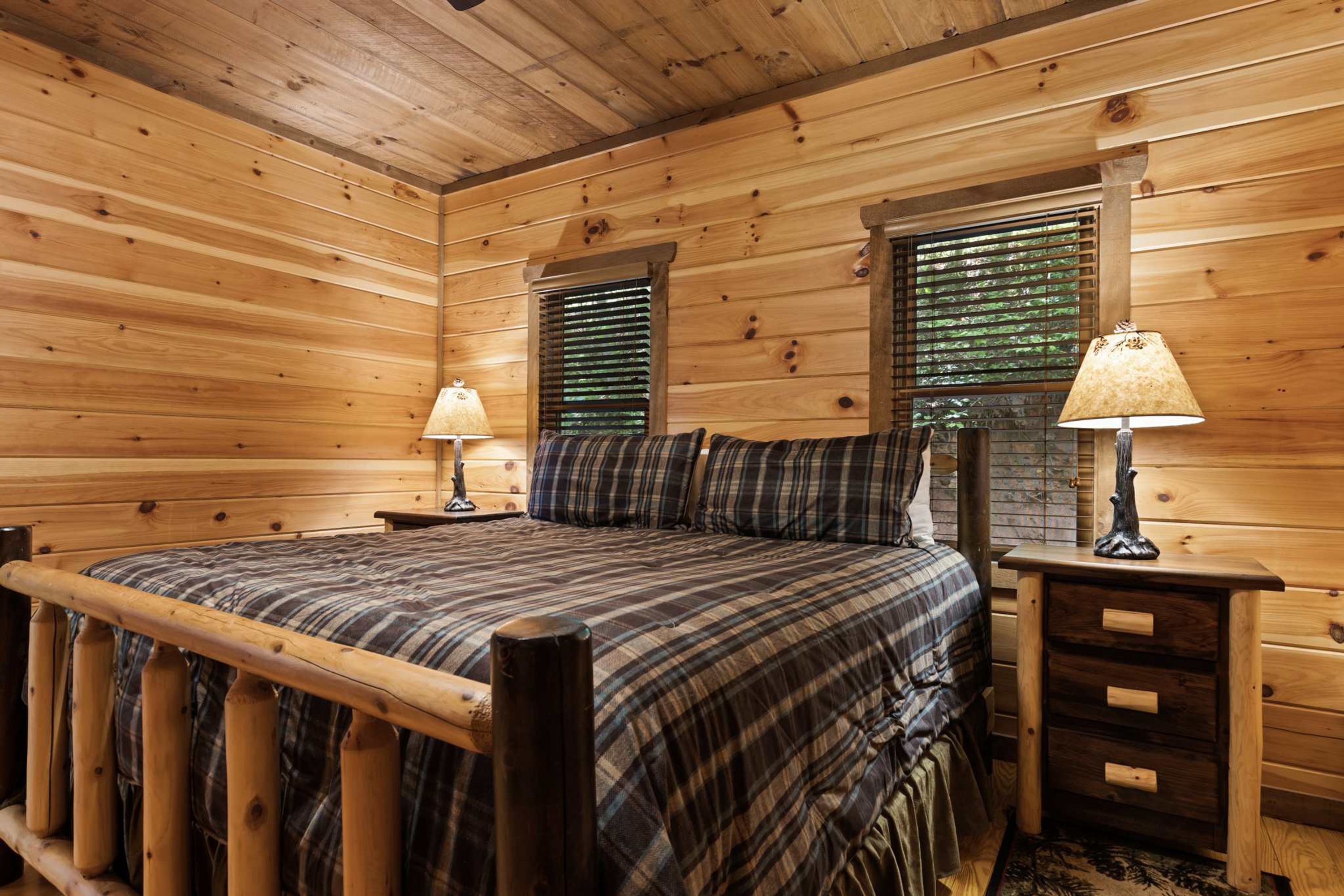 A wooden cabin bedroom with a bed, two nightstands, and a window with blinds.