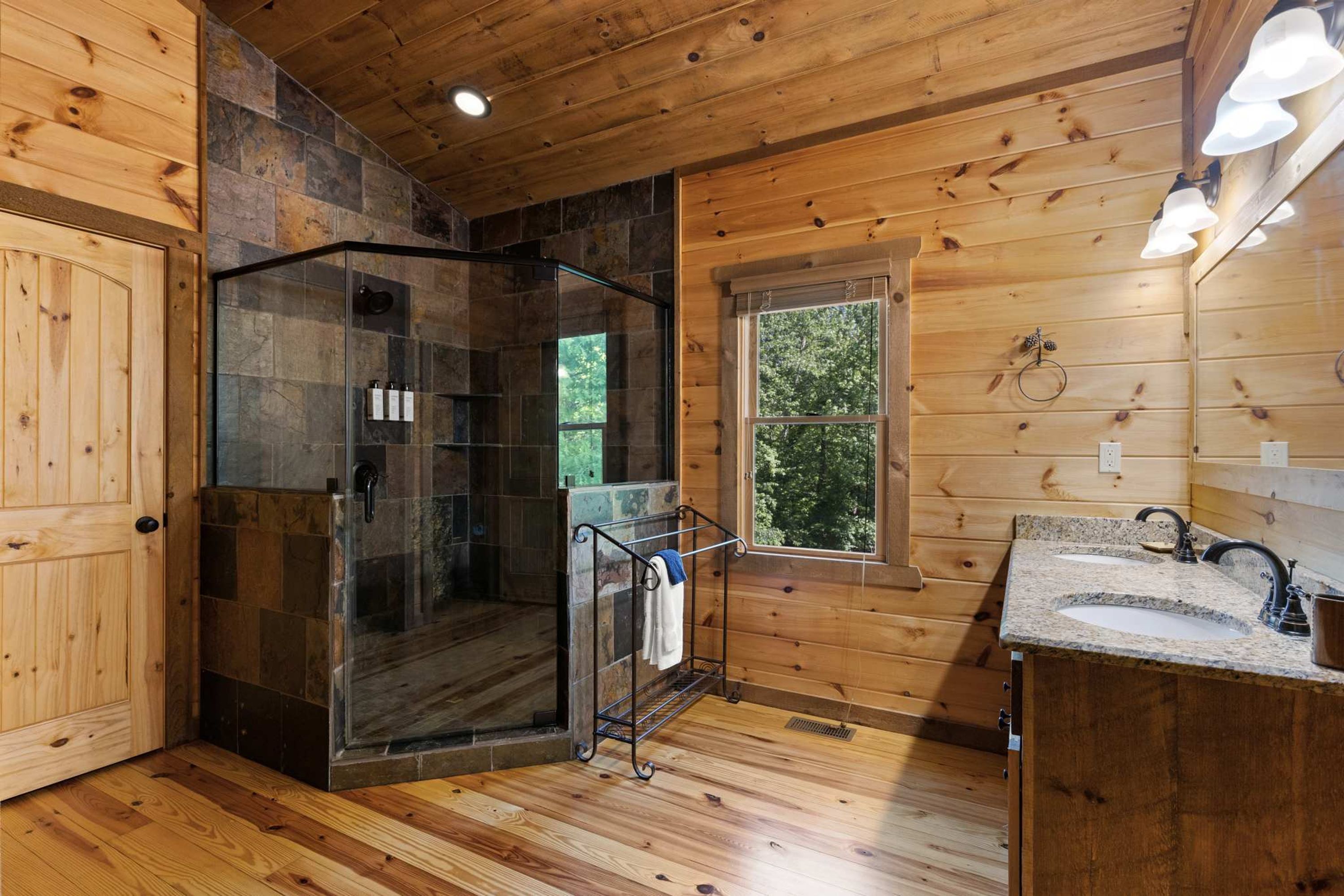 Une salle de bain rustique, avec une cabine en bois, une douche fermée par des parois de verre, une fenêtre et un lavabo doté d’un plan de travail en granit.