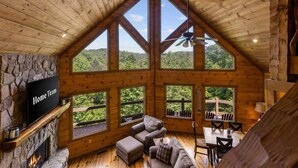A spacious living room with a stone fireplace, a television, and a large window overlooking a forested area. - Mtn Cabin w Hot Tub Game Room Grill Views (Blue Ridge)