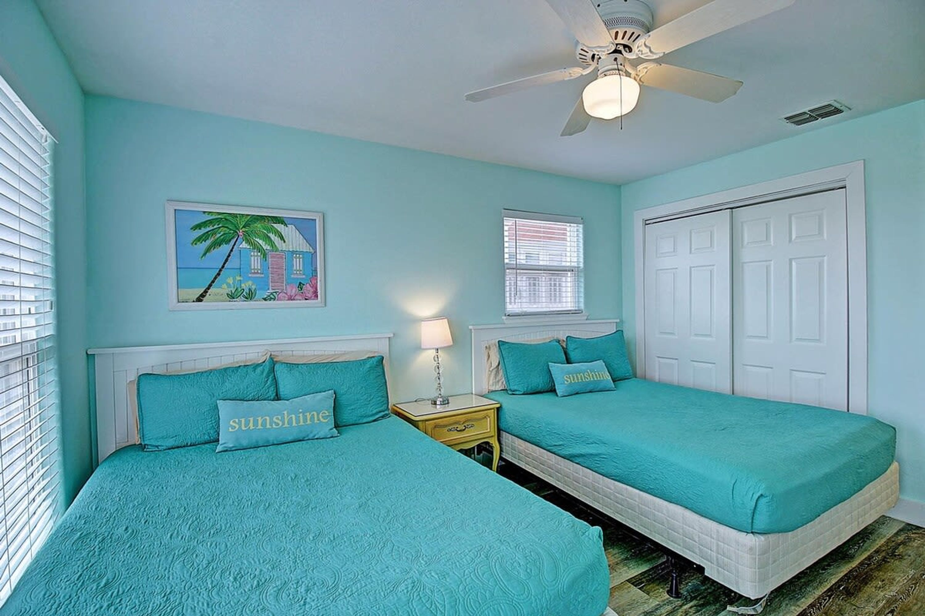 A room with two beds, a ceiling fan, a painting, and a window with blinds.