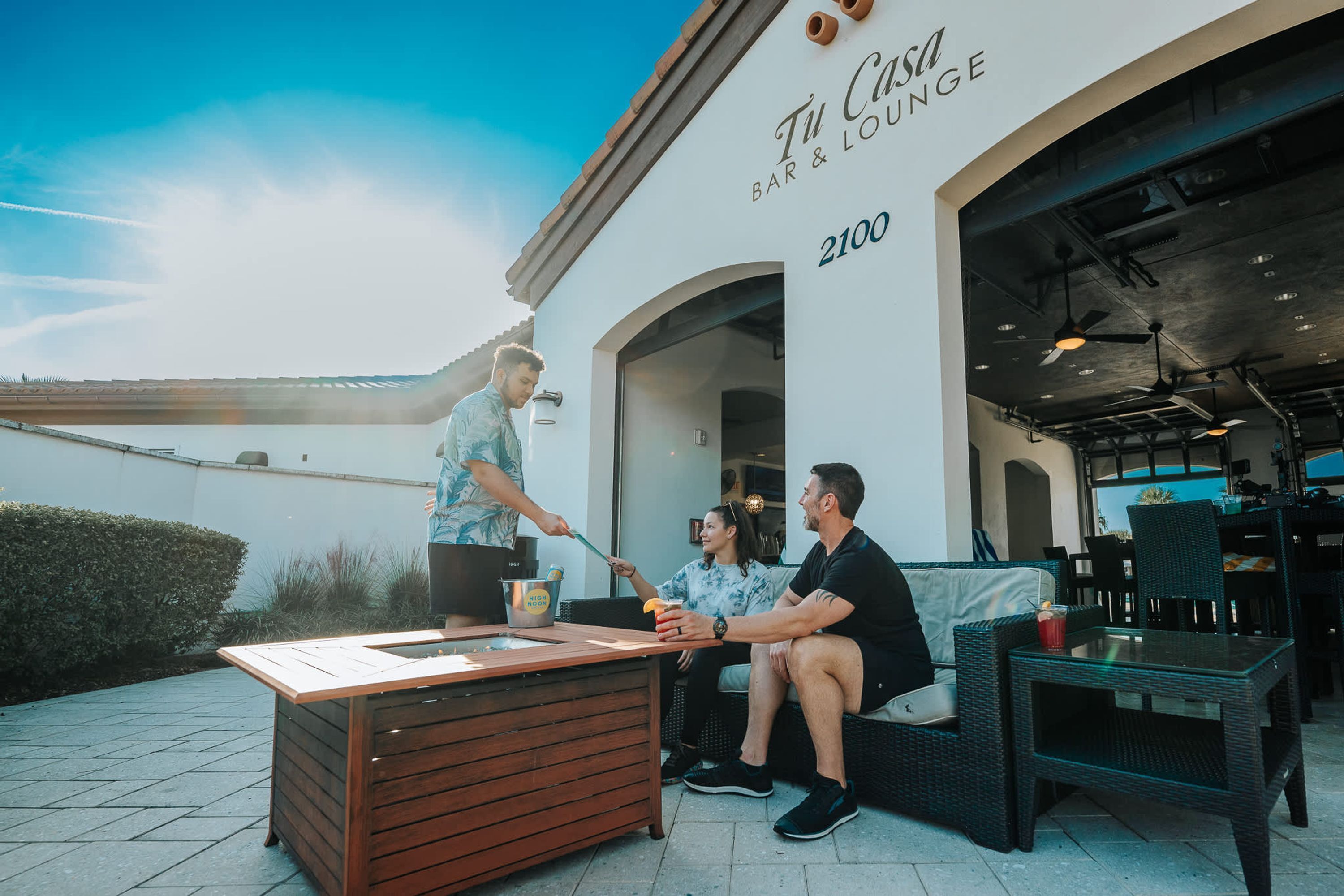 Un espace extérieur aménagé avec un comptoir de bar, des meubles en osier et un bâtiment portant l’enseigne « Tu Casa Bar & Lounge ».