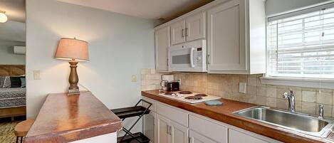 Cocina compacta con armarios blancos, microondas, lavamanos y una barra con taburetes.