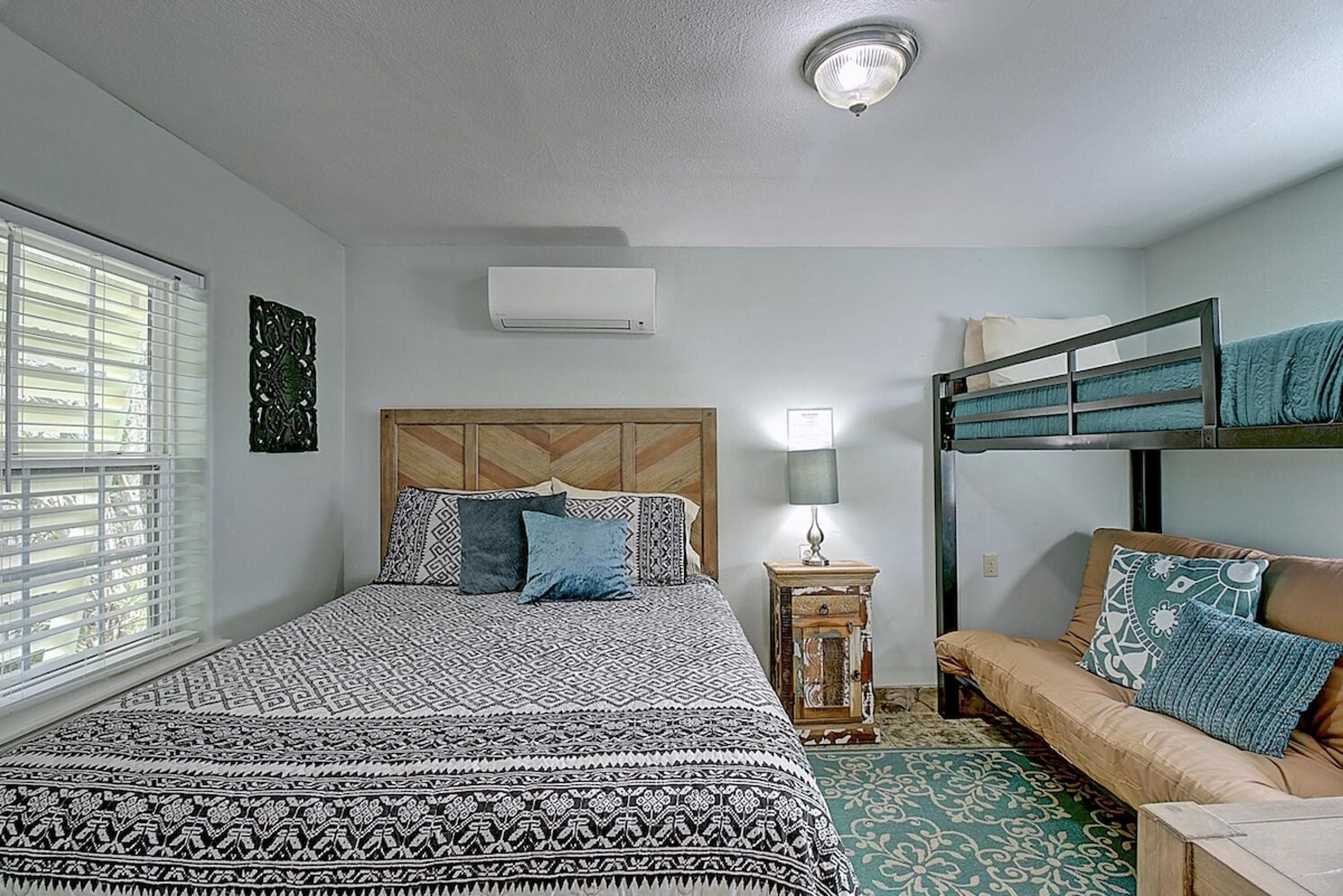A bedroom with a bunk bed, a single bed with patterned bedding, a bedside table with a lamp, and a window with blinds.