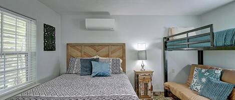 A bedroom with a bunk bed, a single bed with patterned bedding, a bedside table with a lamp, and a window with blinds.