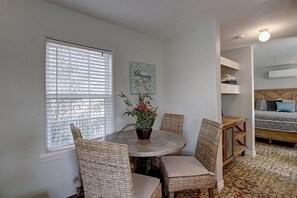 Ein Zimmer mit einem runden Tisch, Korbstühlen, einer Topfpflanze und einem Bett mit gemusterten Betttextilien.