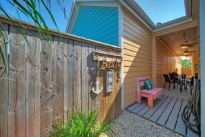 A wooden deck with a bench and a table, a wooden fence with a sign, and a building with a blue roof. - Sparkle Fish Private Breezeway Resort Pool (Port Aransas)