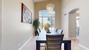 Uma sala de jantar com uma mesa comprida, cadeiras, um lustre e uma janela grande com vista.