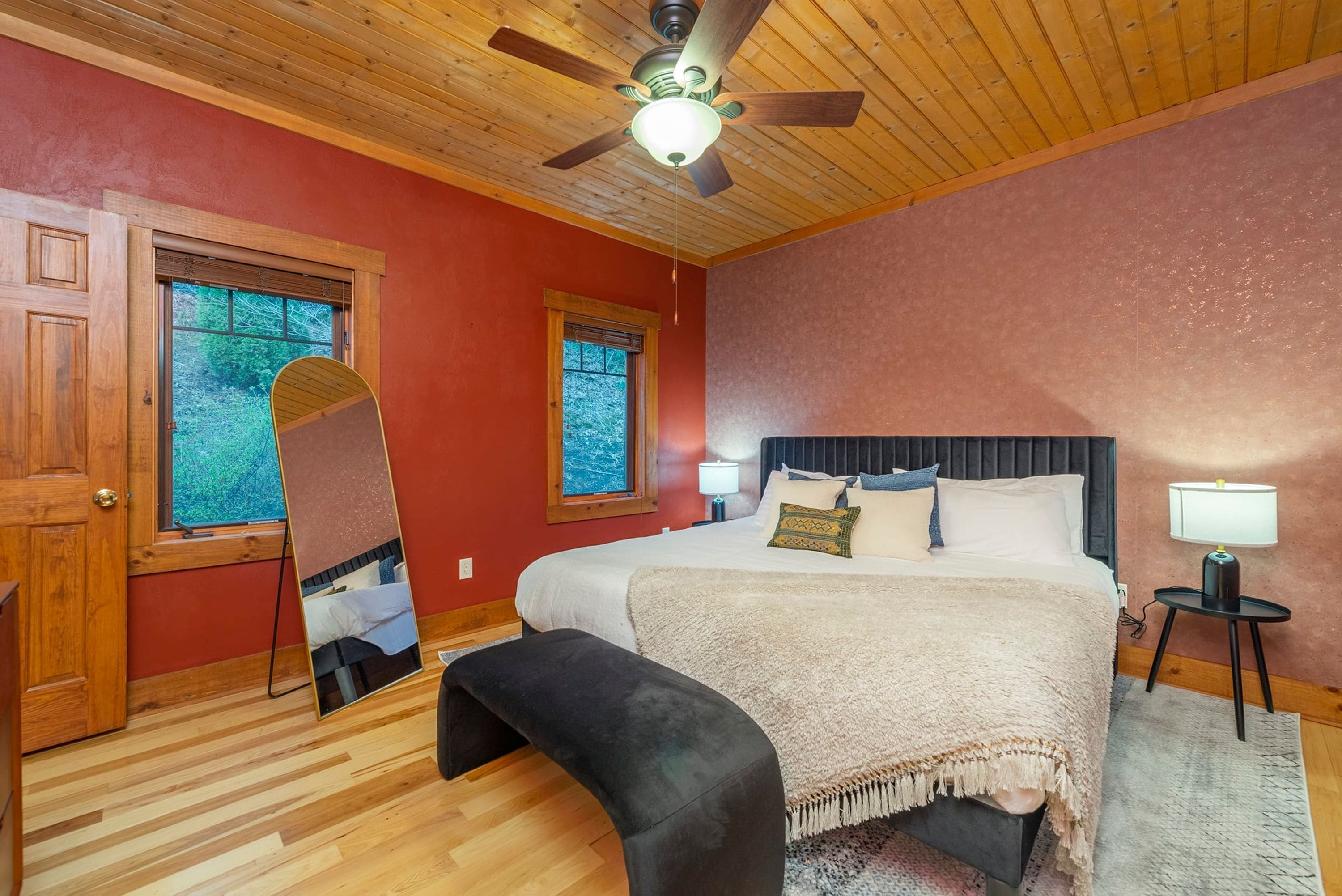 A bedroom with a wooden ceiling, a bed with a dark headboard, two bedside lamps, and a large window with a view of trees.
