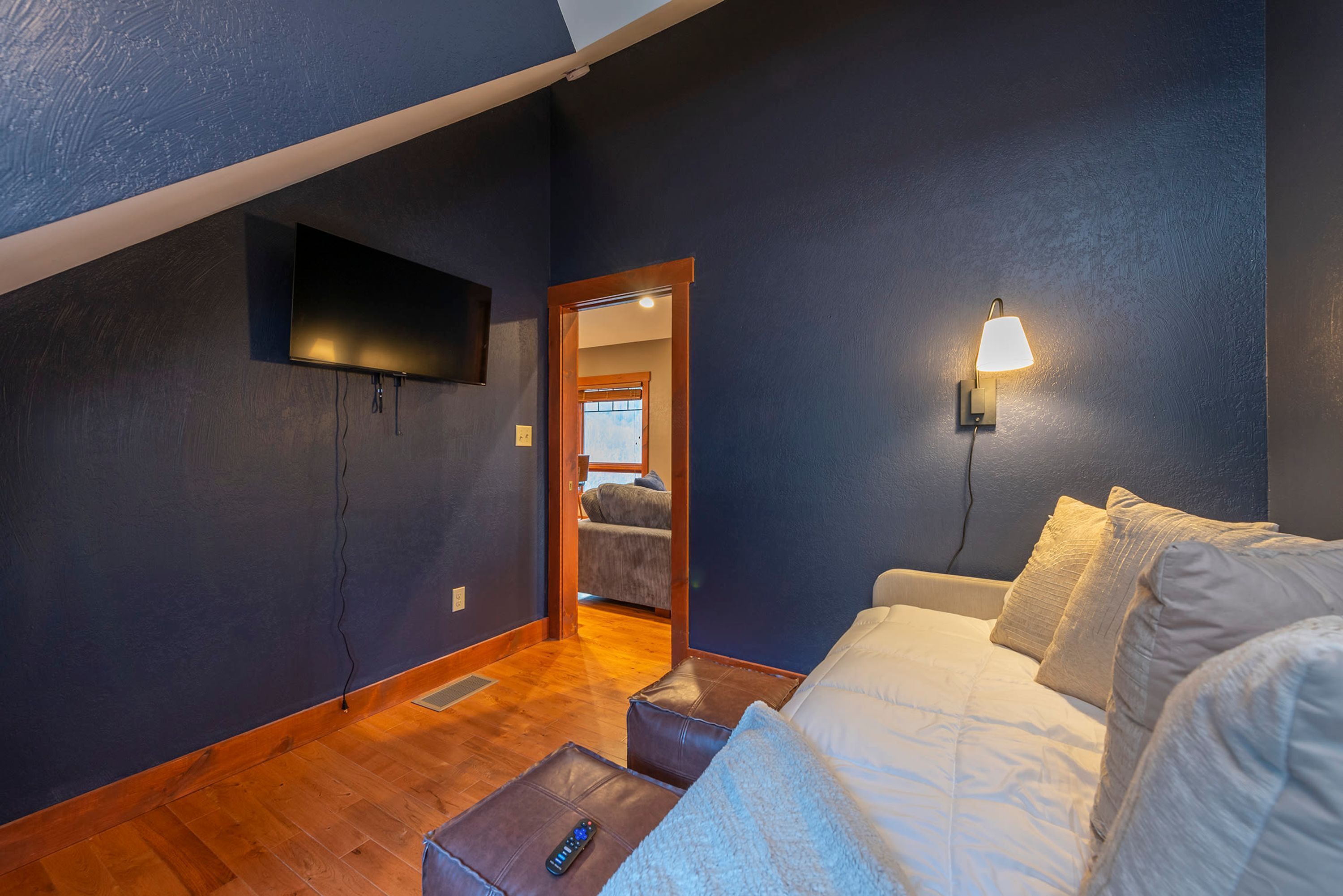 Un dormitorio con una cama, un televisor montado en la pared, una lámpara y un control remoto sobre una mesita.