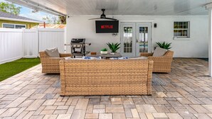 A covered patio area with a central seating arrangement, a ceiling fan, and a mounted TV screen displaying 'NETFLIX'. - Coastin Close Family Fun HTD Pool Outdoor Games (Pinellas Park)