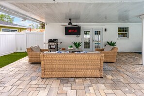 A covered patio area with a central seating arrangement, a ceiling fan, and a mounted TV screen displaying 'NETFLIX'. - Coastin Close Family Fun HTD Pool Outdoor Games (Pinellas Park)