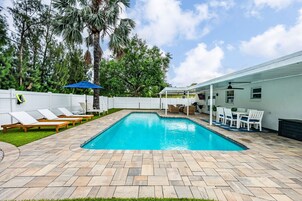 Una piscina rectangular con solárium de baldosas, rodeada por una valla blanca y un área de descanso al aire libre con mobiliario blanco.