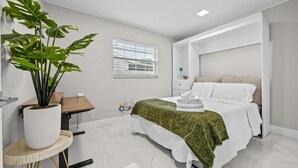 A modern bedroom with a bed, a bedside table, a lamp, a potted plant, and a window with blinds. - Coastin Close Family Fun HTD Pool Outdoor Games (Pinellas Park)