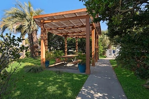 A wooden pergola with a table and chairs, surrounded by lush greenery and a pathway. - Steps to the Beach Pool Courts Family Fun (Destin)