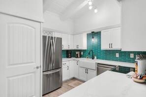 A modern kitchen with white cabinetry, a stainless steel refrigerator, a coffee maker, and a teal backsplash. - Steps to the Beach Pool Courts Family Fun (Destin)