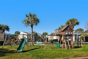 Un parc infantil amb tobogan i cadira de balanceig, envoltat de palmeres i una caseta de joc de fusta.